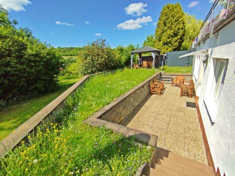 Un beau jardin avec de l'herbe verte et des fleurs colorées. À l'arrière-plan, on peut voir une terrasse et un pavillon.