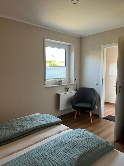 Une chambre simple avec deux lits et une chaise dans le coin. La pièce a une teinte claire et une fenêtre donnant sur l'extérieur.