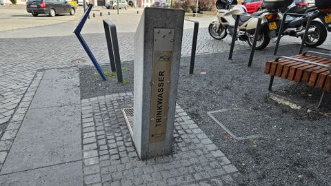 Une source d'eau potable en béton se trouve sur une place. En arrière-plan, des motos et des bancs sont visibles.