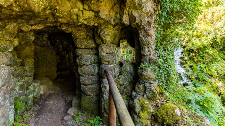 Een rotsachtige, in de rotsen uitgehouwen ingang van een kleine grottengalerij met een pad voorzien van leuningen, omgeven door mos en dicht groen; rechts stort een waterval tussen de rotsen naar beneden.