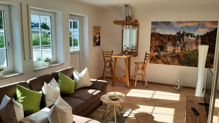 Ein helles Wohnzimmer mit einem gemütlichen Sofa und bunten Kissen. An der Wand hängt ein Bild von einer Burg, und es gibt einen kleinen Tisch mit Stühlen.