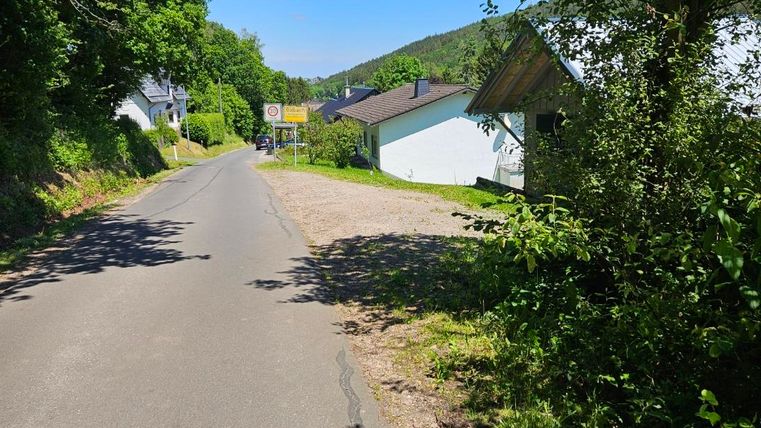 A quiet street, surrounded by trees and green nature. On the left side, there are some houses and in the background, you can see gentle hills.