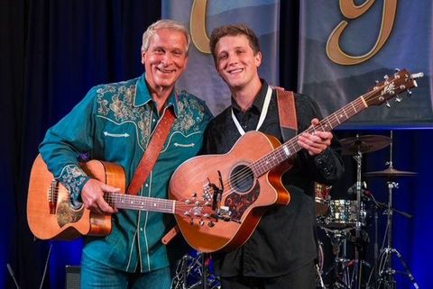 Two musicians are standing on a stage and smiling at the camera. Both are holding acoustic guitars, while a band is visible in the background.
