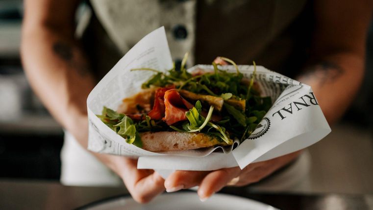 Een vers bereid sandwich met ham en sla, ingepakt in papier. Het wordt vastgehouden door een persoon die aan een tafel staat.