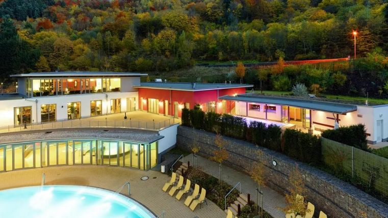 Ein modernes Wellnesshotel mit einem einladenden Außenpool. Die Umgebung ist von grünen Hügeln und buntem Laub geprägt.
