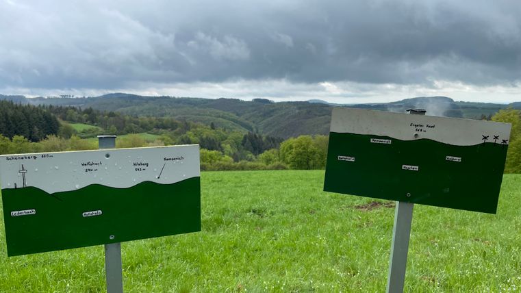 Zwei Schilder auf einer Wiese zeigen die Namen und Höhen von Bergen in der Eifel. Im Hintergrund sind bewaldete Hügel und ein bewölkter Himmel zu sehen.
