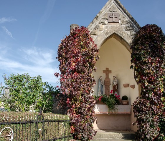 Une petite chapelle à Wolsfeld, entourée de plantes grimpantes rouges. A l'intérieur, on peut voir des statues religieuses. Le ciel est clair et bleu., © Tourist-Information Bitburger Land