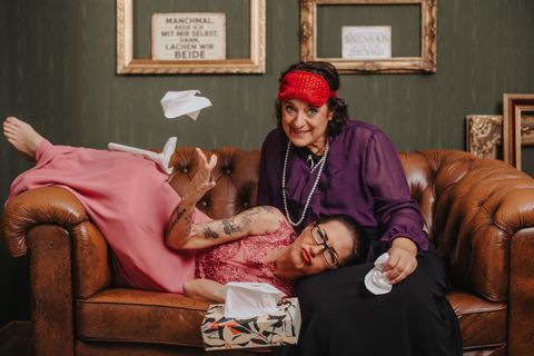 Two women are sitting on a leather couch. One woman, wearing a sleep mask, is throwing tissues into the air, while the other woman, who is lying on her lap, has a tearful expression and is holding a tissue in her hand. There is a box of tissues on the couch.