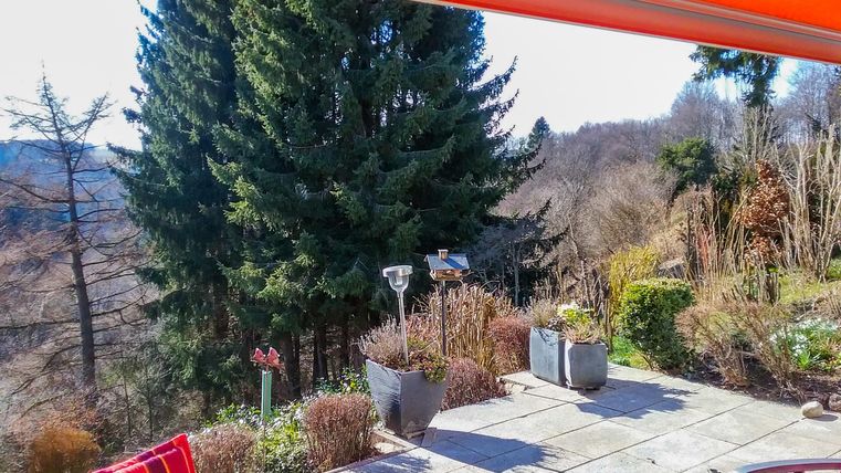 Une terrasse avec des chaises rouges et une vue sur un paysage forestier. Le soleil brille et le ciel est bleu.