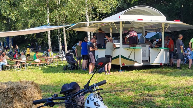 Een drukke snackbar in de natuur, omringd door mensen die eten en praten. Op de voorgrond staat een fiets met helm.