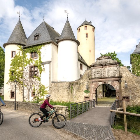 Zwei Radfahrer fahren an der historischen Burg Rittersdorf vorbei, die von grünen Bäumen und einem klaren Himmel umgeben ist., © Eifel-Tourismus GmbH/Dominik Ketz