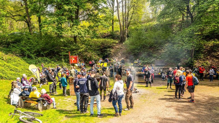 A lively gathering of people in a forest. Some are standing and chatting, while others are sitting by the campfire and relaxing.