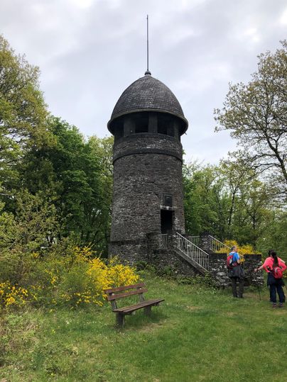 A historic stone observation tower, surrounded by trees and green vegetation. In the foreground, there is a bench, and hikers are on their way.