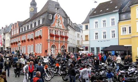 Een levendig stadsplein met veel motorfietsen en een grote menigte. Op de achtergrond zijn historische gebouwen in verschillende kleuren te zien.