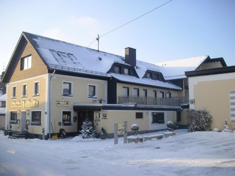 Een gezellig huis in de sneeuw, omgeven door een winterlandschap. De lucht is helder en de zon schijnt zachtjes op de besneeuwde omgeving.