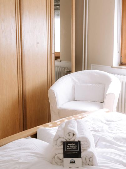 A cozy bedroom with a white armchair and a wooden wardrobe. Freshly folded towels are lying on the bed.