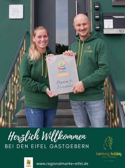 A warm welcome to the Eifel hosts. A couple proudly holds a certificate in front of a modern building.