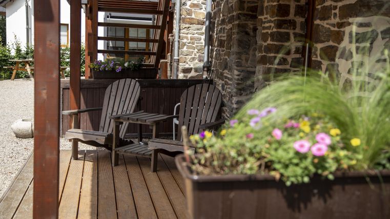 Une terrasse cosy avec deux chaises et des plantes fleuries. Le sol est en bois et en arrière-plan, on peut voir des murs en pierre.