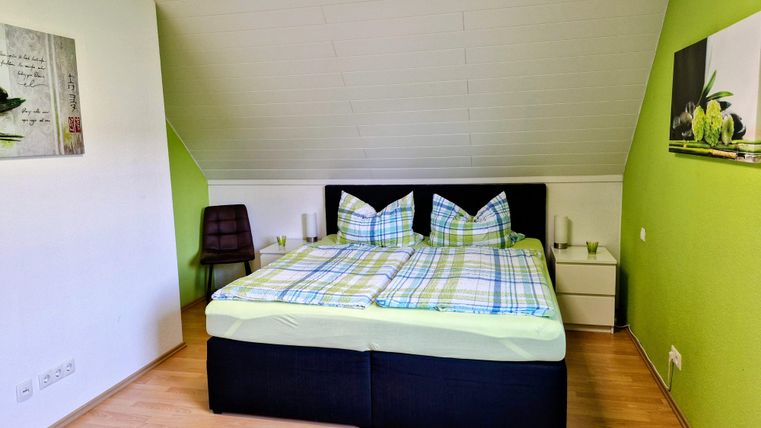 Een moderne slaapkamer met een groot bed en kleurrijke kussens. De muren zijn geschilderd in een frisse groentint en er is een stoel en decoratieve afbeeldingen.