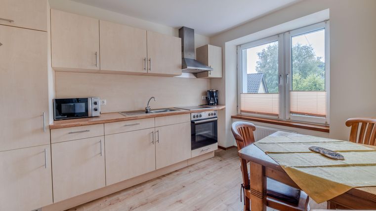 Een moderne keuken met lichte houten kasten en een groot raam. Er is een eettafel met stoelen en verschillende keukengerei.