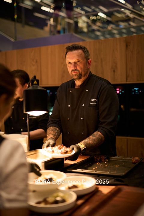 Een kok staat in een moderne keuken en presenteert gerechten. Op de achtergrond zijn tafels en een stijlvolle inrichting te zien.