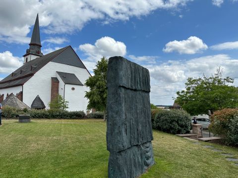 Kirche mit spitzem Turm und moderner Skulptur im Vordergrund, umgeben von grünem Rasen und Bäumen.