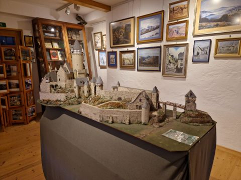 Une vue modèle détaillée d'un château est posée sur une table. En arrière-plan, des cadres photo sont visibles sur le mur.