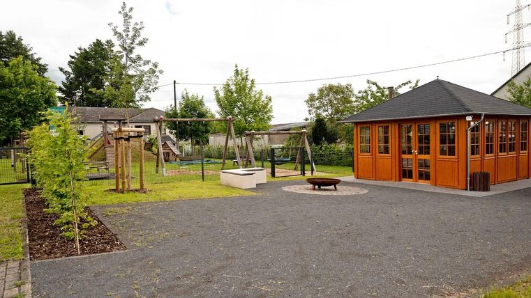 Ein Spielplatz mit Schaukeln und einem Holzhaus. Die grüne Umgebung ist mit Bäumen und einer gepflasterten Fläche gestaltet.