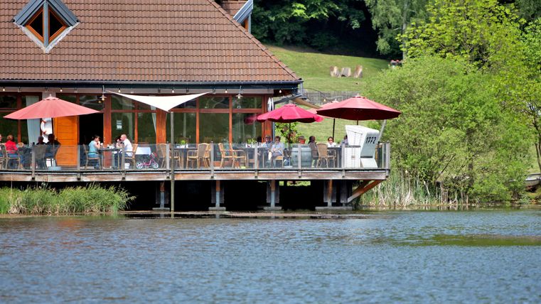 Ein gemütliches Restaurant am See mit roten Schirmen. Die Umgebung ist grün und einladend.