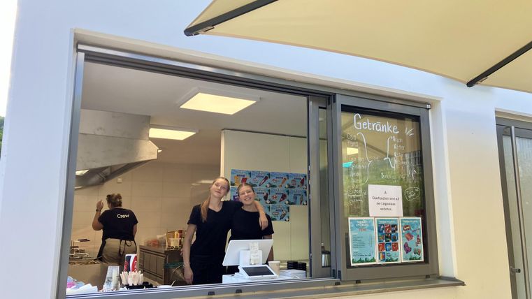 Deux femmes se tiennent à un comptoir et sourient. En arrière-plan, on peut voir des appareils de cuisine et des affiches colorées.