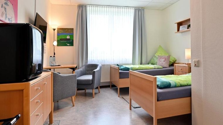 Ein einfaches Zimmer mit zwei Einzelbetten und einem Fenster, das viel Tageslicht lässt. Es gibt einen Sessel, einen Fernseher und einen Schrank für persönlichen Bedarf.