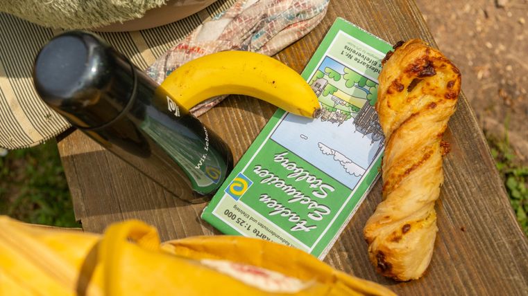 Een picknick met een broodje, een banaan en een thermoskan op een houten tafel. De omgeving lijkt zonnig en ontspannend te zijn.