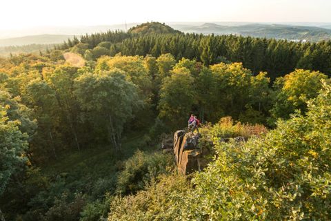 Two people are standing on a rock with a view of a forest and hills in the background.