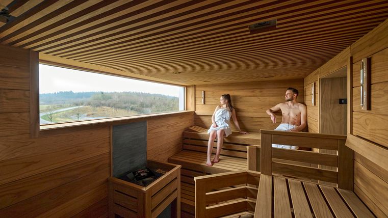 Een moderne sauna met houten bekleding en grote ramen. Twee personen ontspannen op de banken en genieten van het uitzicht.