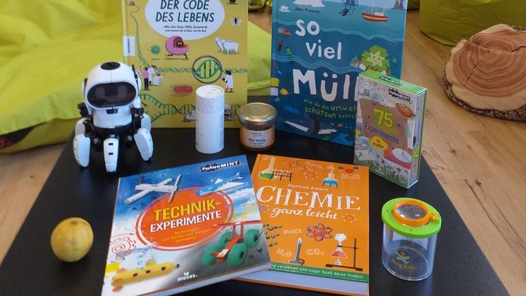 Ein Tisch mit verschiedenen Büchern, darunter Titel über Technik und Chemie. Außerdem sind ein kleiner Roboter, ein Glas und ein Apfel zu sehen.