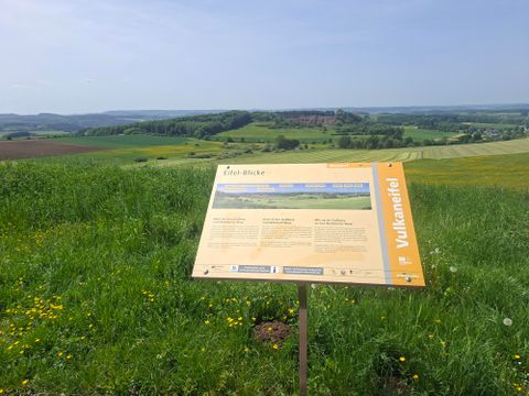 Un panneau d'information se dresse sur une pelouse verte avec une vue sur le paysage vallonné. À l'arrière-plan, on peut voir des forêts et des champs.