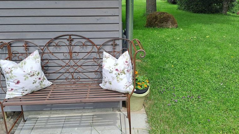 A metal bench with two decorative cushions is placed on a paved area. In the background, a well-maintained lawn can be seen.