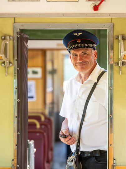 Ein Zugschaffner steht in der Tür eines Waggons und lächelt. Der Innenraum ist hell und die Sitzgelegenheiten sind gut sichtbar.