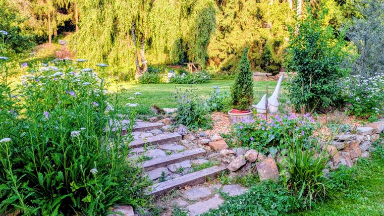 Ein schöner Garten mit bunten Blumen und einer Treppe aus Steinen. Im Hintergrund sind grüne Bäume und Sträucher zu sehen.