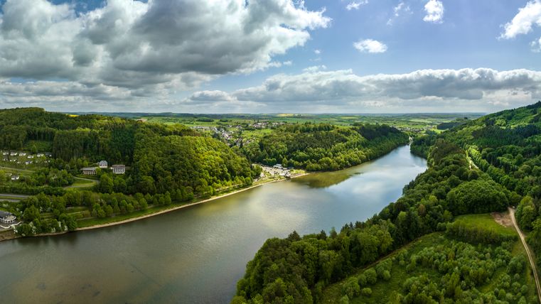 Aerial view of the Bitburg reservoir in Biersdorf am See, surrounded by green forests and hills under a cloudy sky.