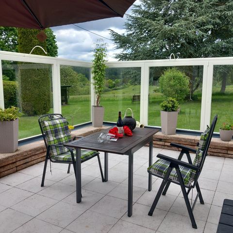 Een gezellige eetruimte buiten met een tafel en twee stoelen. Op de achtergrond zijn groene planten en een rustige landschap te zien.