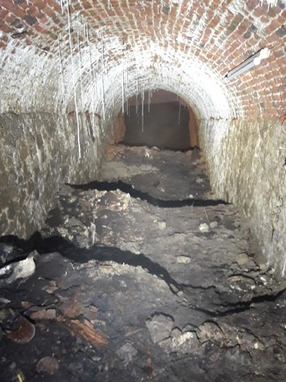 Een donkere tunnel met rode bakstenen muren en een oneffen vloer. Er liggen hier en daar wat latten en vuil op de vloer.