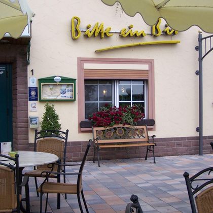 Een gezellig terras met tafels en stoelen voor een klein restaurant. Het bord van het restaurant toont de naam „Graag een biertje“ en bloeiende planten sieren het raam.