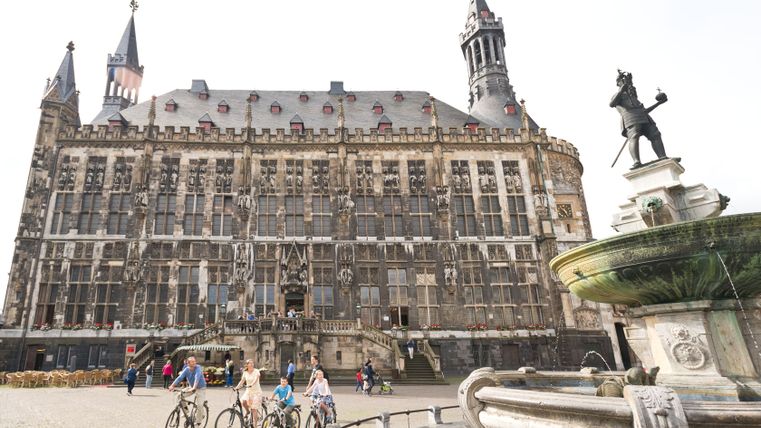 Hôtel de ville d'Aix-la-Chapelle avec une fontaine et des cyclistes au premier plan.
