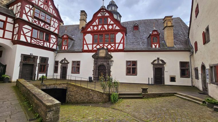 De schilderachtige binnenhof van kasteel Bürresheim met historische vakwerkhuizen. De bodem is met stenen bestraat, en grijze wolken trekken voorbij aan de lucht.