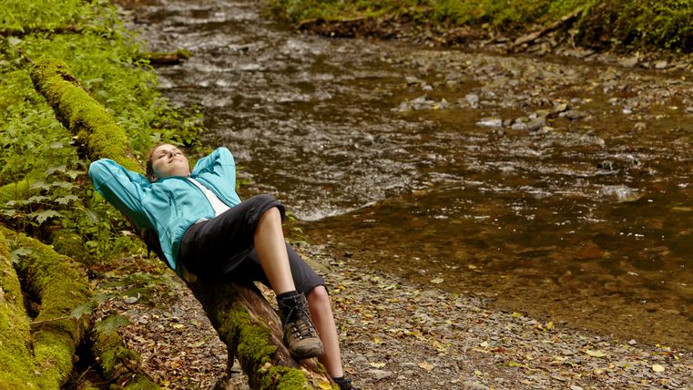Wandelaar leunt ontspannen tegen een met mos bedekte boomstam aan de oever van een beek.