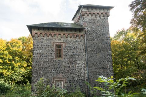 An old stone building with two towers stands in a green environment. Colorful trees in the background give the scene an autumn atmosphere.