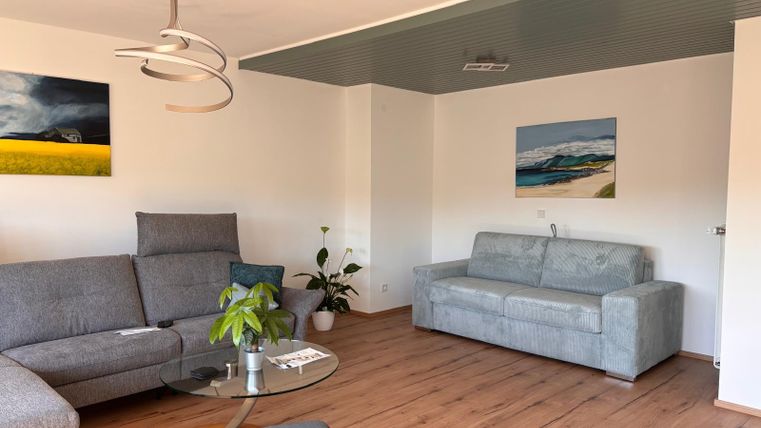 A modern living room with a gray couch and a glass table. Various pictures hang on the walls, and a plant stands in the corner.