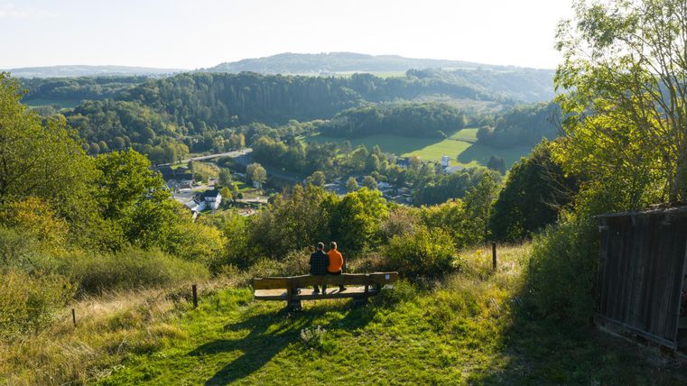A view of a picturesque landscape with gentle hills and plenty of greenery. Two people are sitting on a bench and enjoying the view.