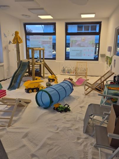 Ein Spielzimmer mit Sandboden und verschiedenen Spielgeräten. Spielzeug wie Autos und ein Tunnel sind ebenfalls vorhanden.